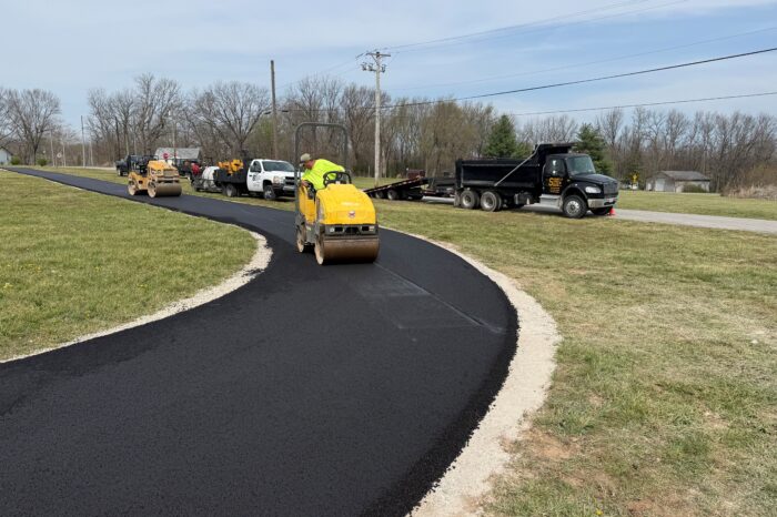 Workers paving asphalt road with nearby trucks and equipment.