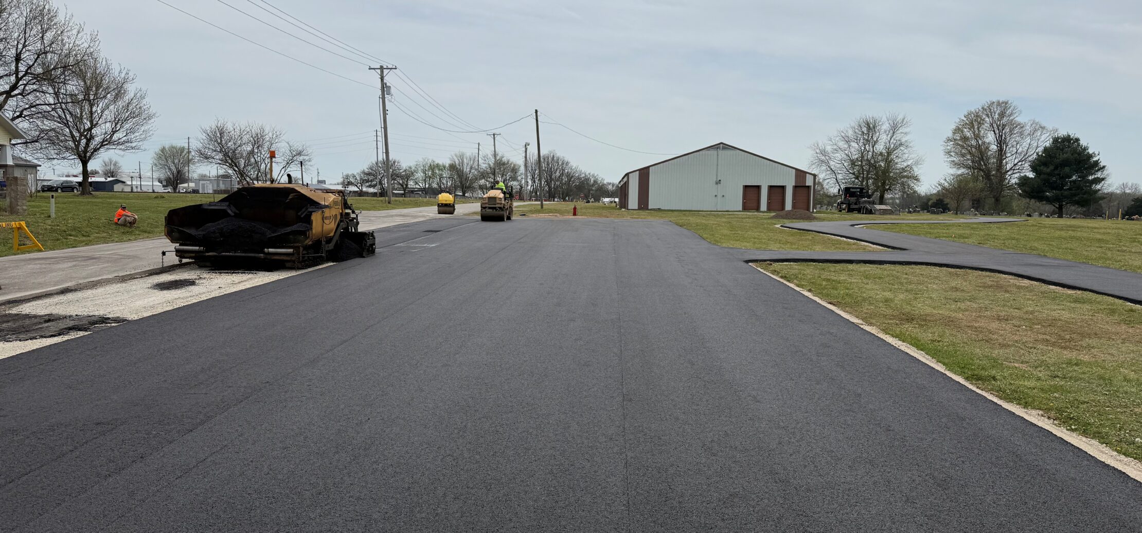 Workers paving asphalt road with nearby trucks and equipment.