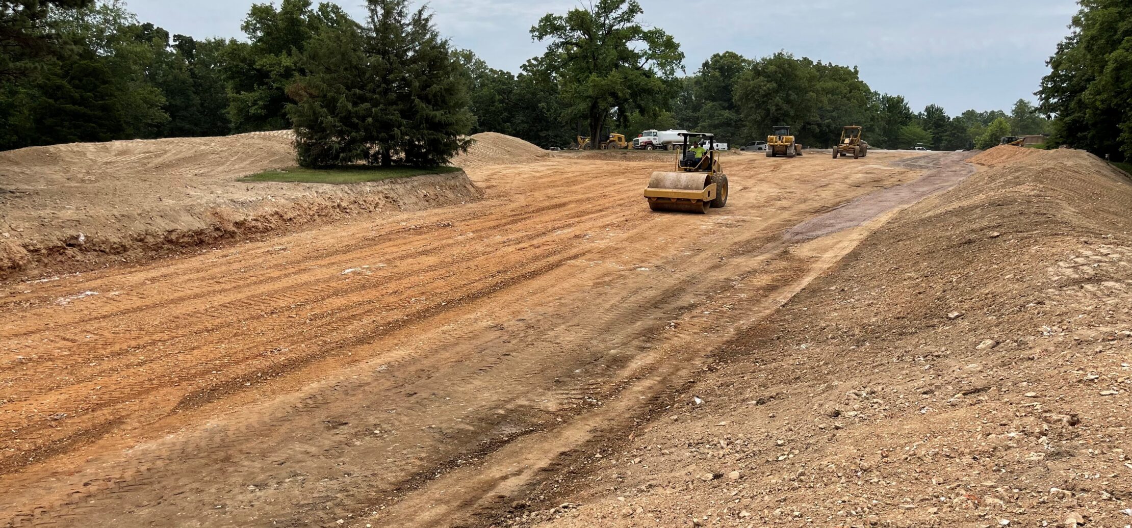 Workers pave new asphalt road with machinery, trees nearby.