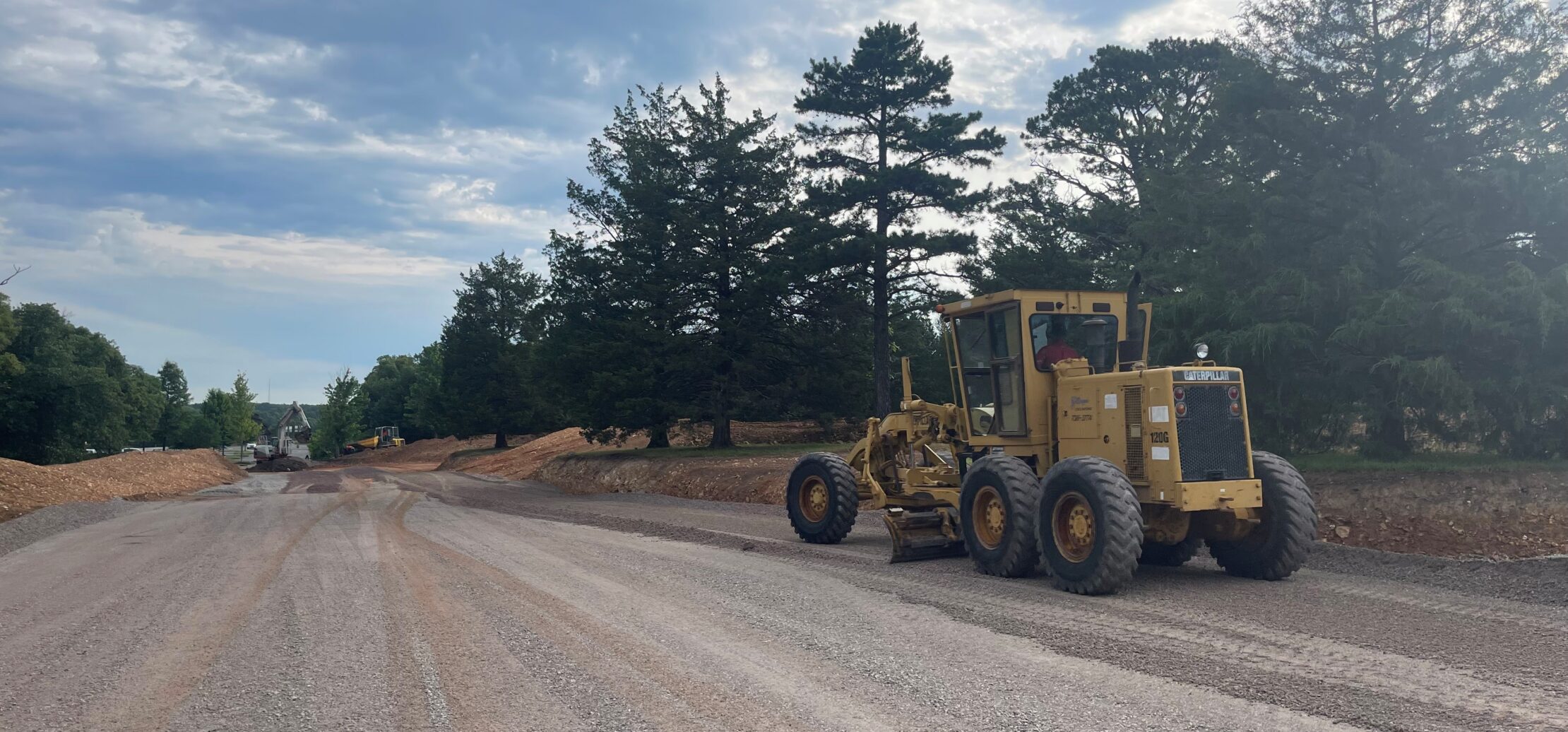 Workers pave new asphalt road with machinery, trees nearby.