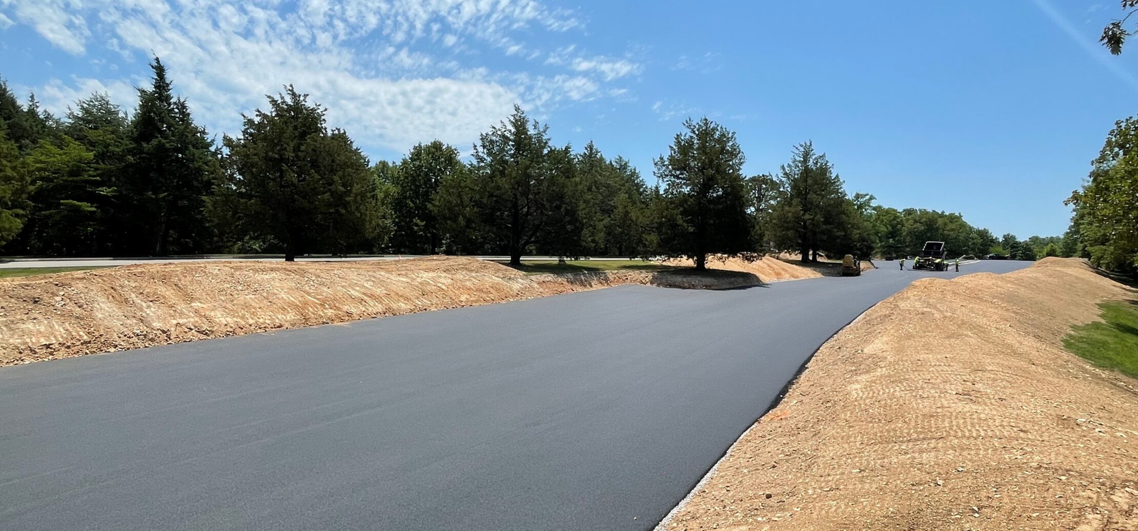 Workers pave new asphalt road with machinery, trees nearby.