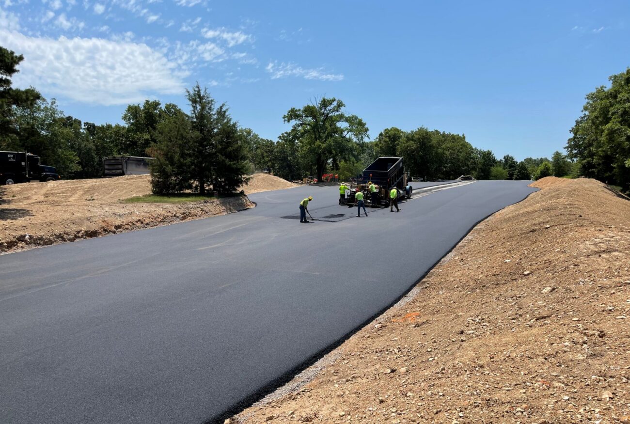 Workers pave new asphalt road with machinery, trees nearby.