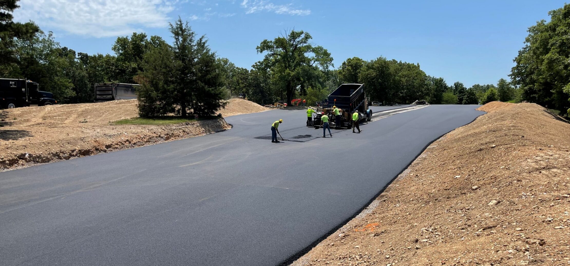 Workers pave new asphalt road with machinery, trees nearby.