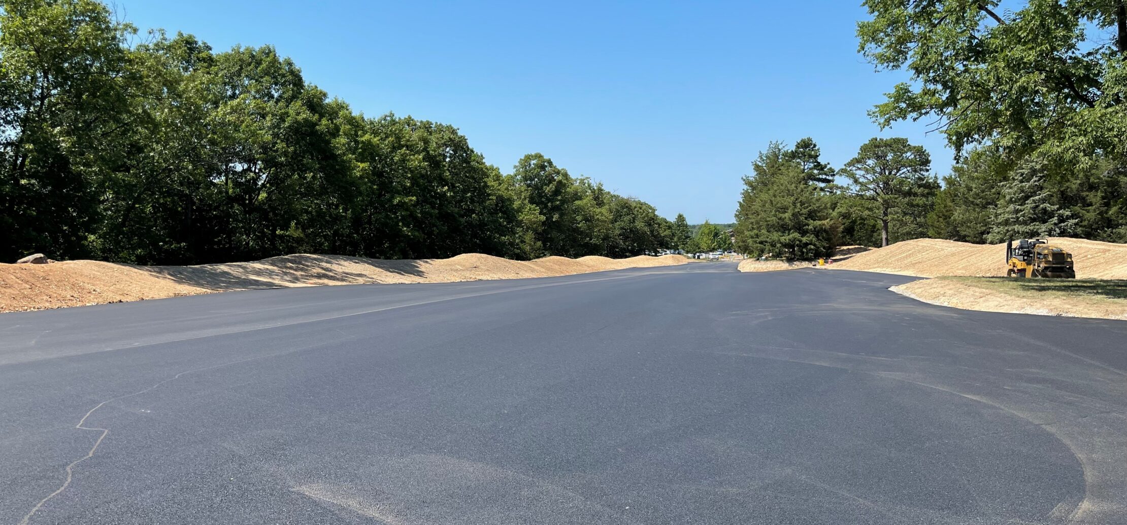 Workers pave new asphalt road with machinery, trees nearby.