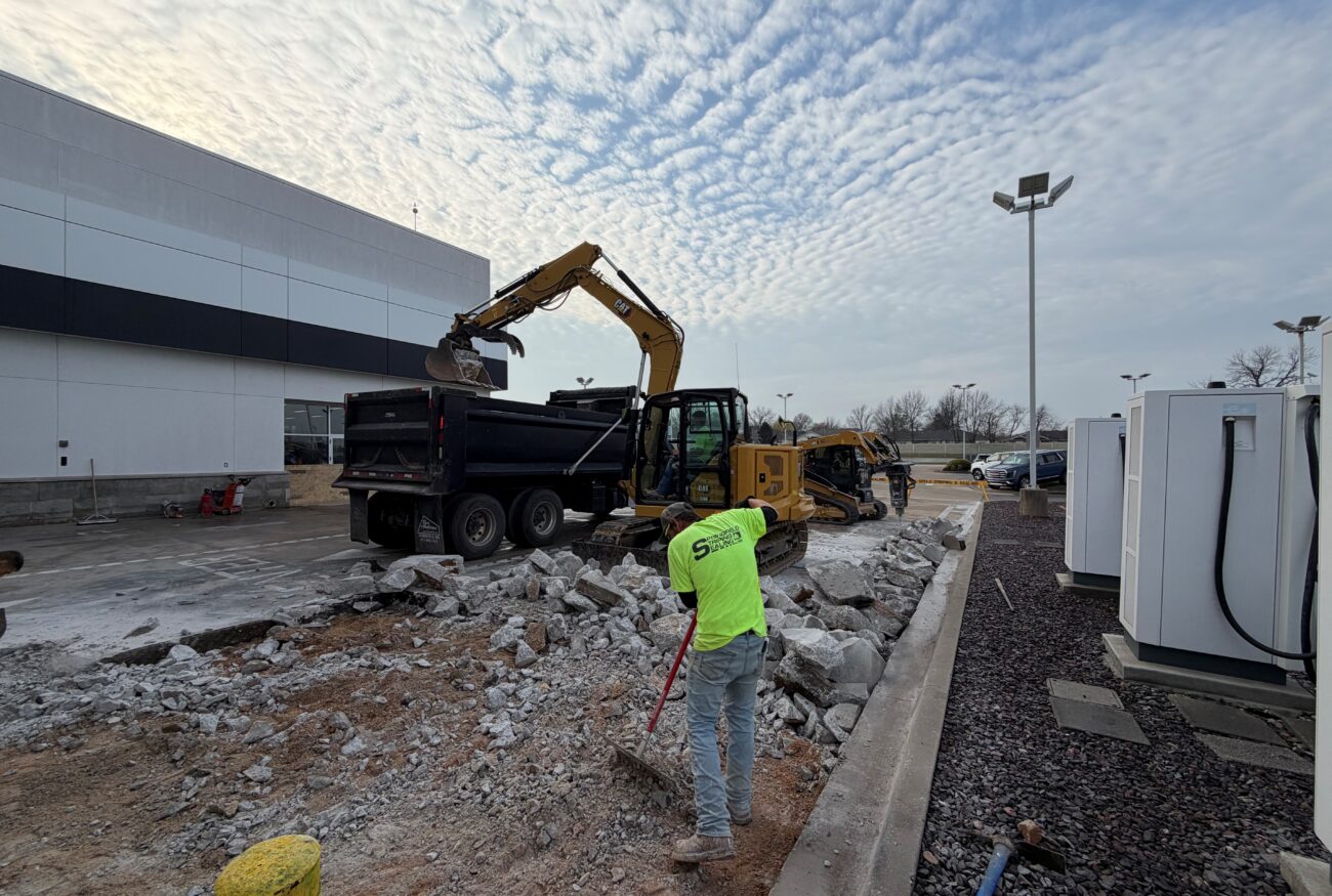 Workers remove concrete rubble with machinery outside a commercial building.