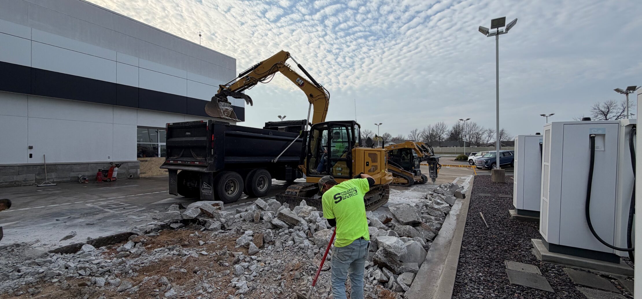 Workers remove concrete rubble with machinery outside a commercial building.
