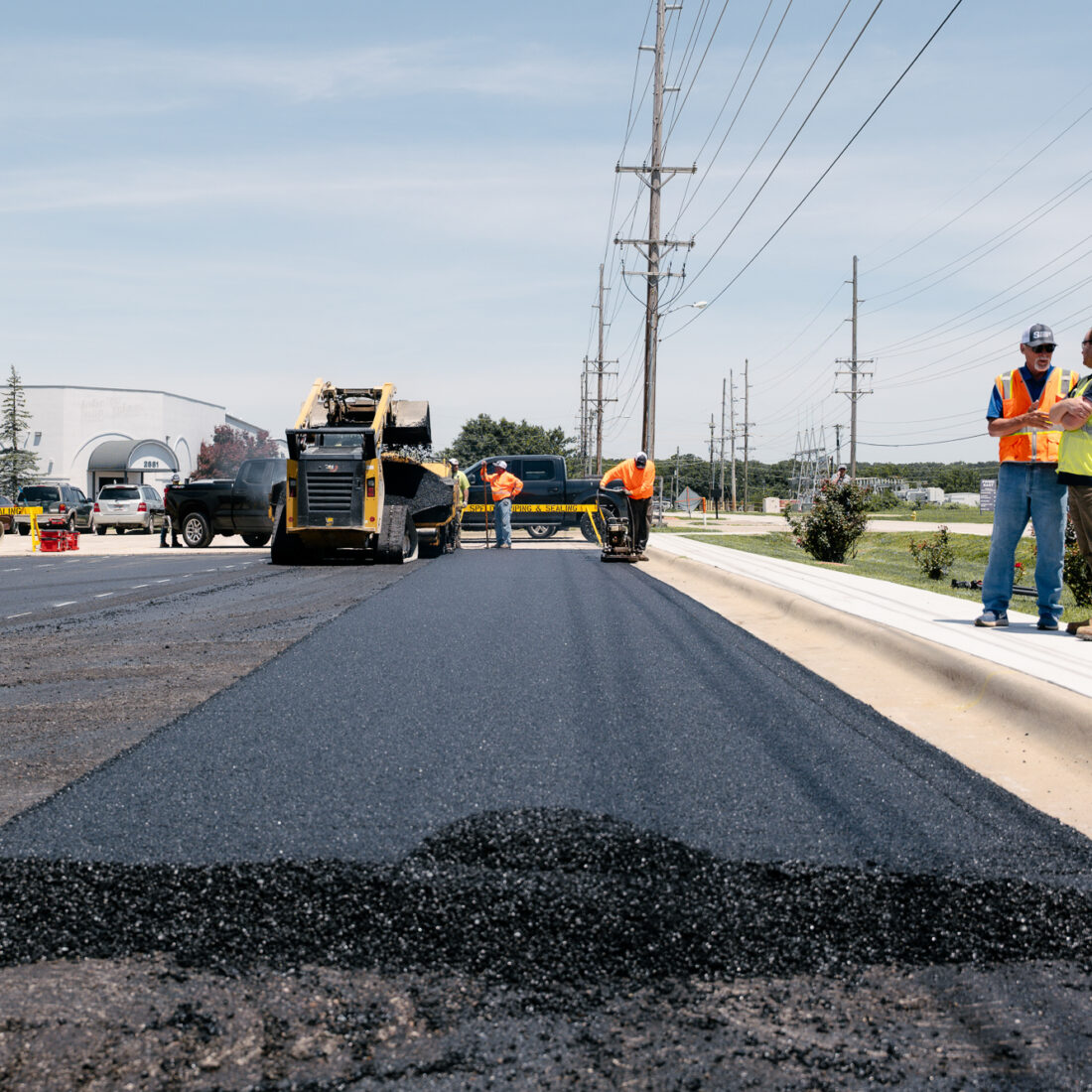 Workers pave road with machinery on sunny day.