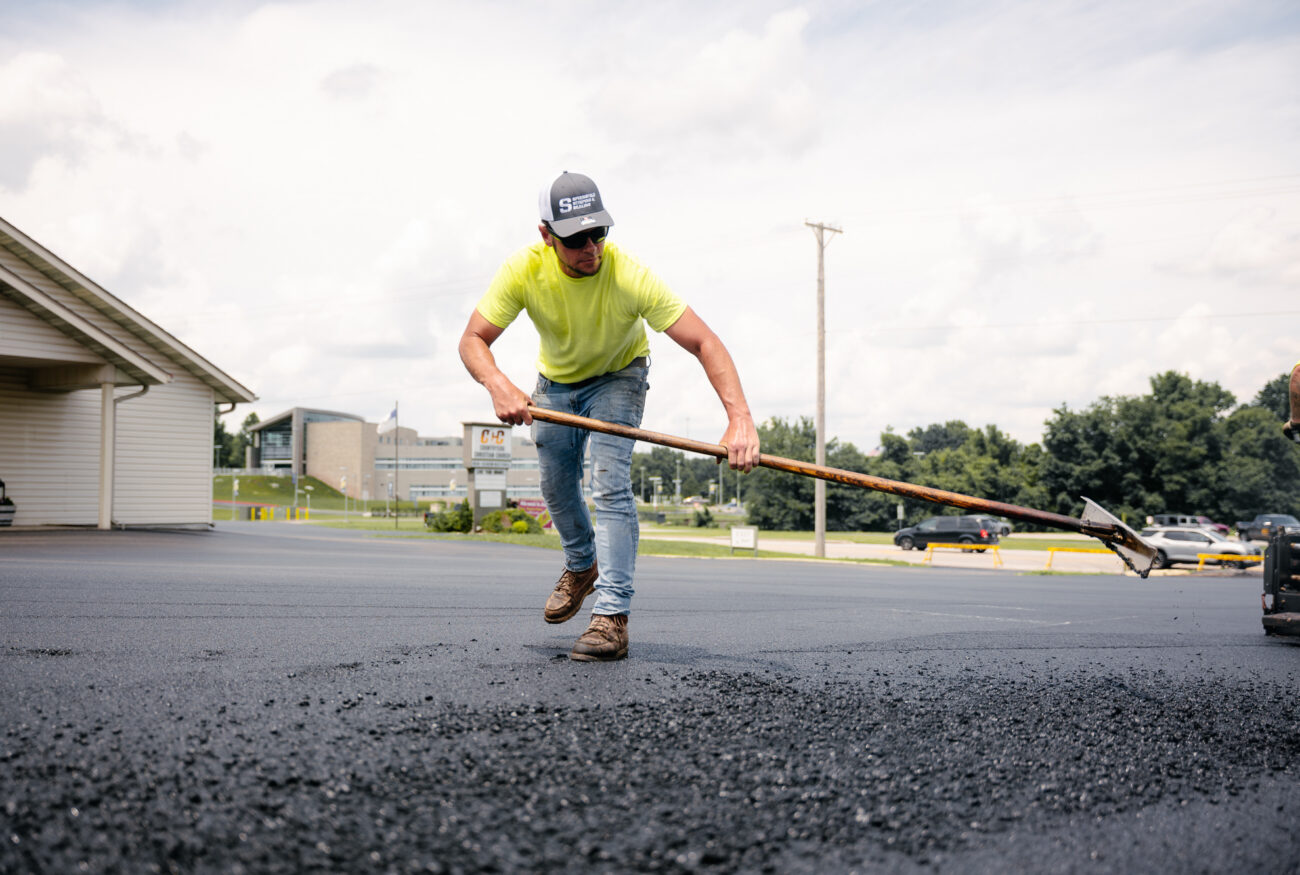 Worker in yellow spreading asphalt on parking lot.
