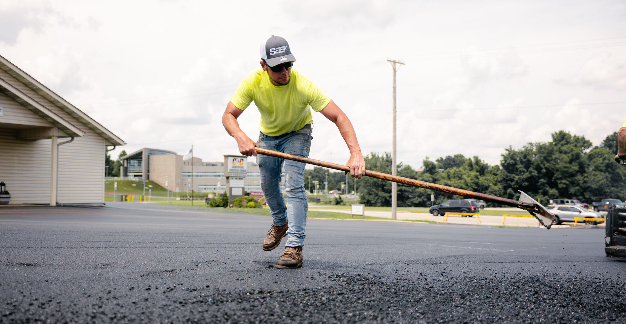 Worker in yellow spreading asphalt on parking lot.