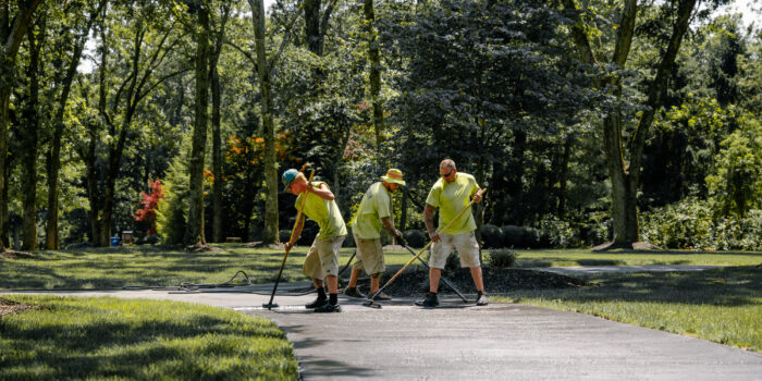 Three workers in yellow clean a park walkway with tools.