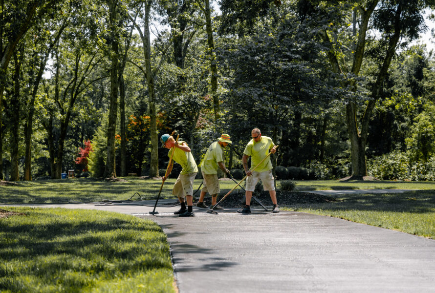 Three workers in yellow clean a park walkway with tools.