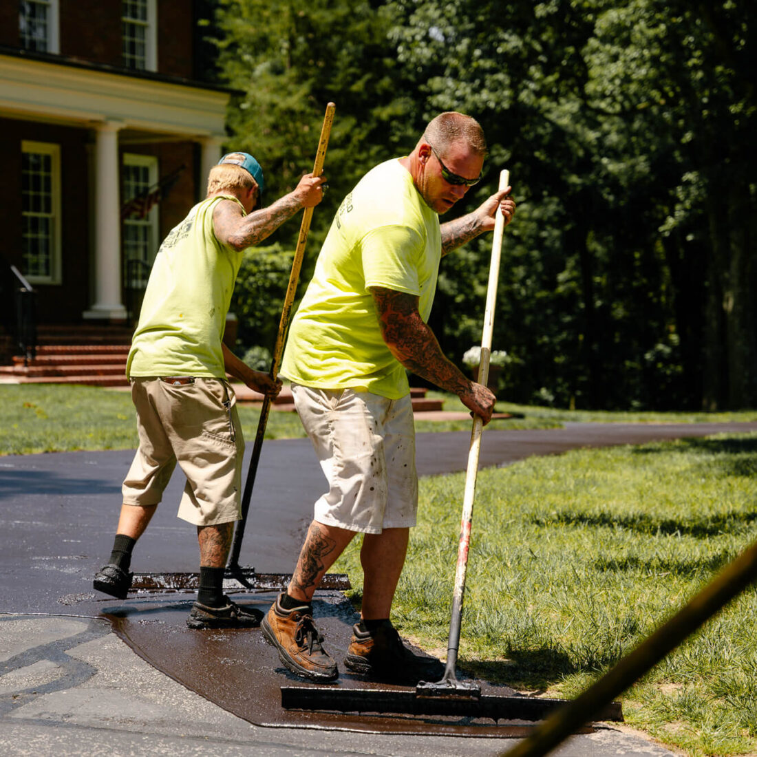 Two workers spread asphalt on a driveway near a house.