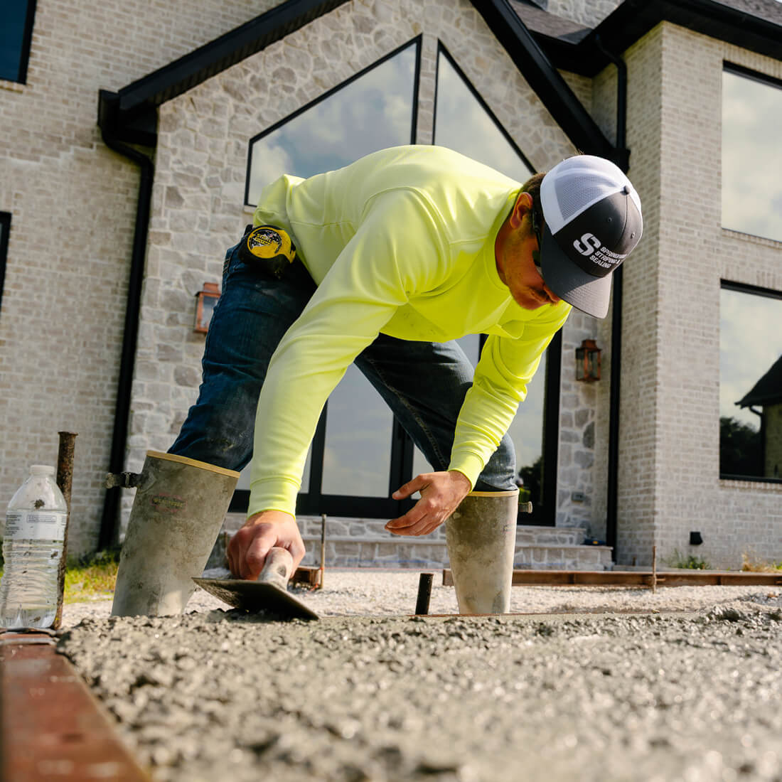 Worker in yellow shirt smooths concrete by stone house.