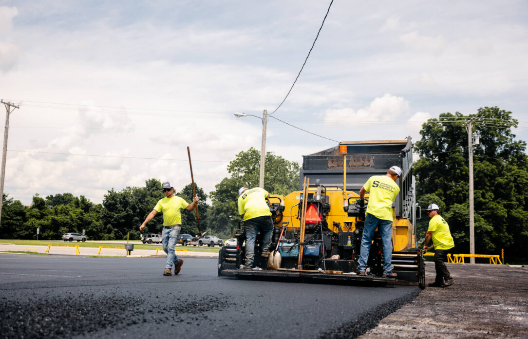 Construction workers repave road with machinery; sunny, cars nearby.