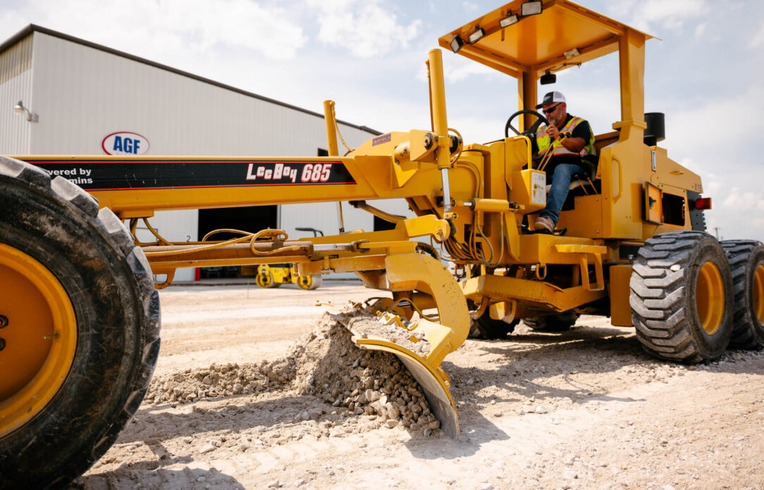 Worker operates yellow grader near metal building, leveling gravel.