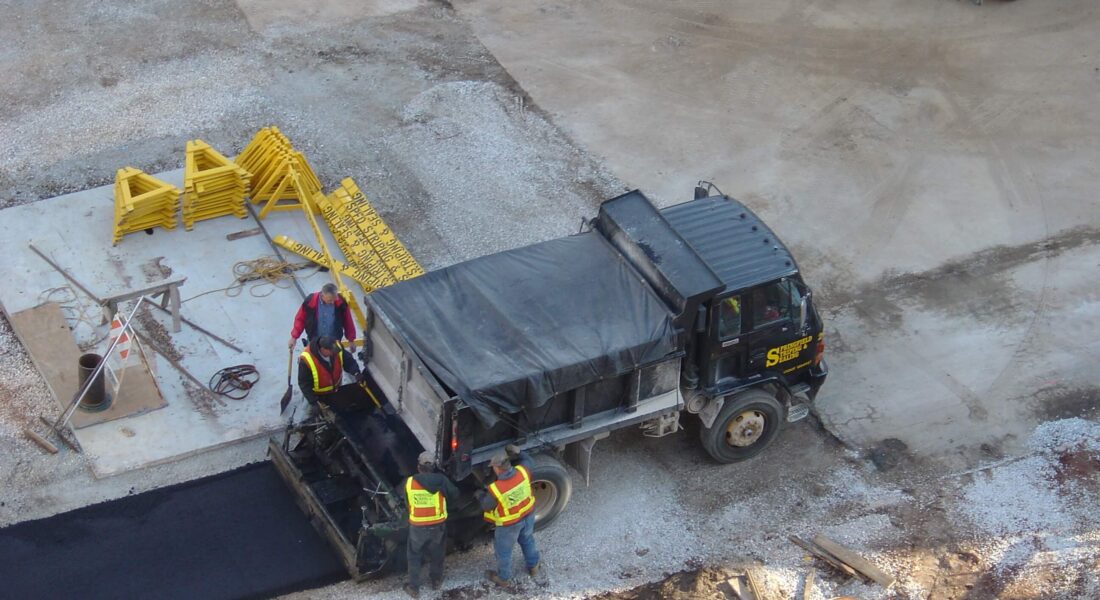 Construction workers pave road with asphalt near truck and barricades.