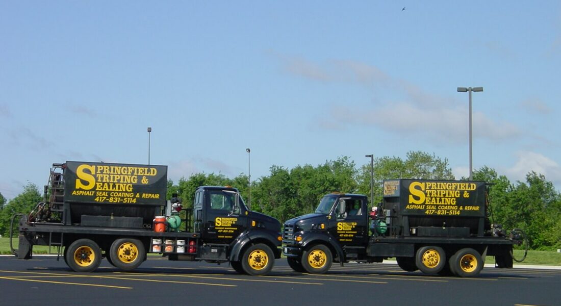 Two black trucks with yellow lettering on striped asphalt lot.