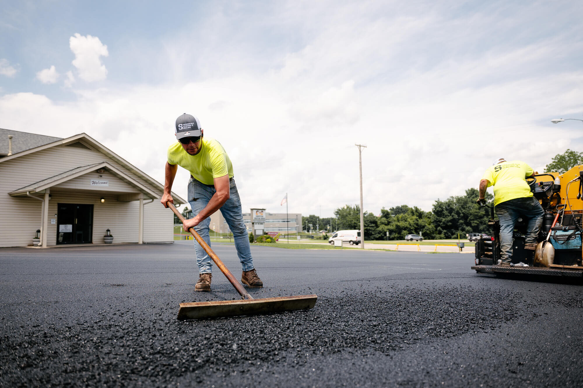 Worker rakes asphalt; another drives paving machine nearby.