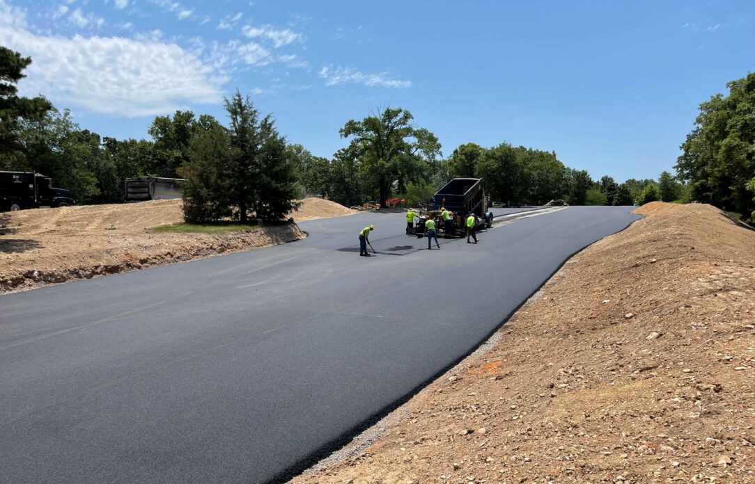 Workers pave asphalt road; equipment, dirt, and trees nearby.