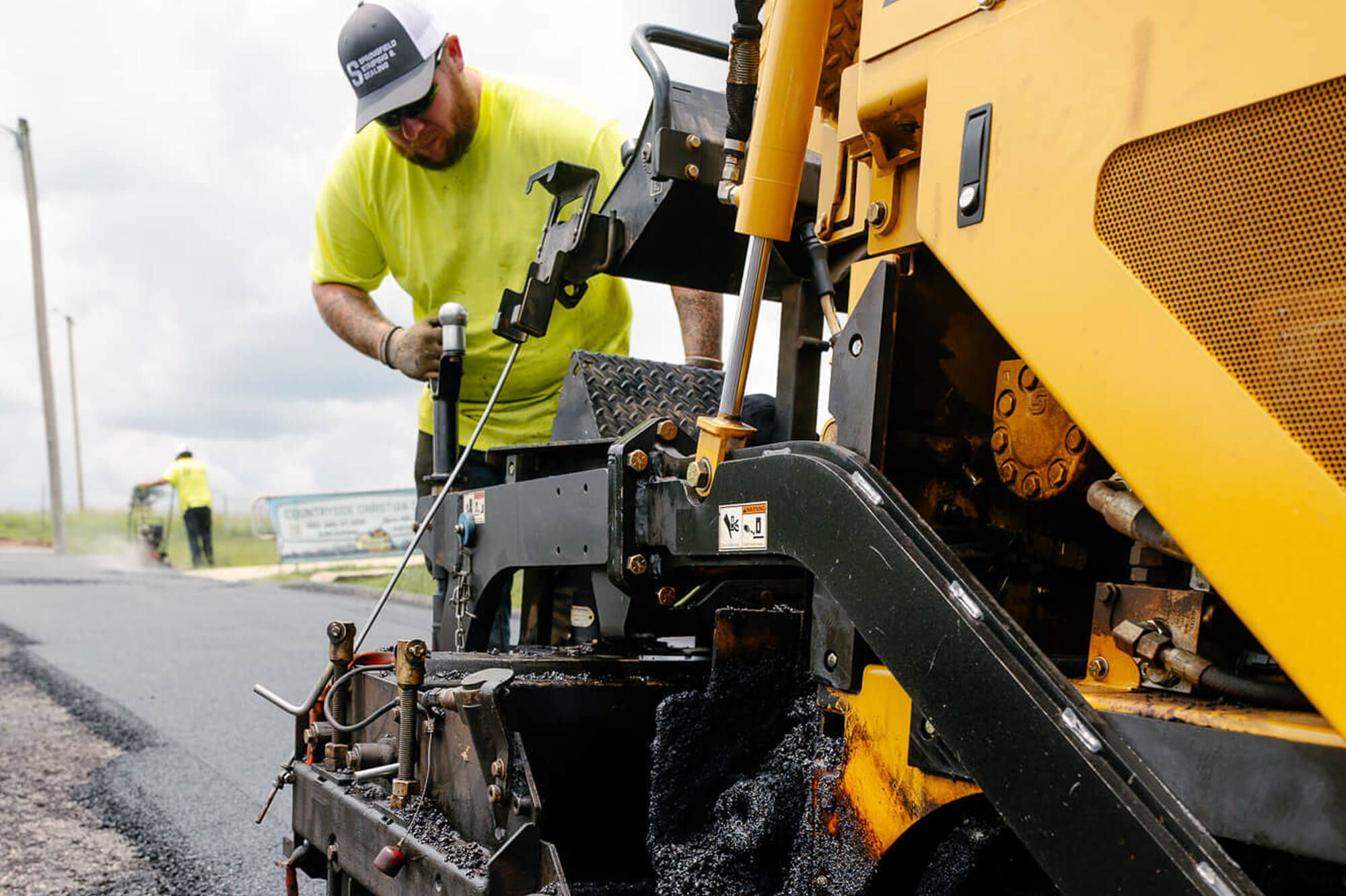 Worker in yellow shirt operates asphalt paver on cloudy day.
