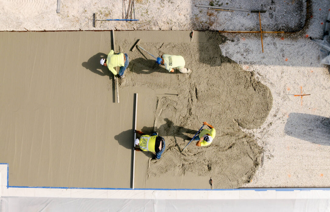 Four workers smooth wet concrete on a rectangular slab.