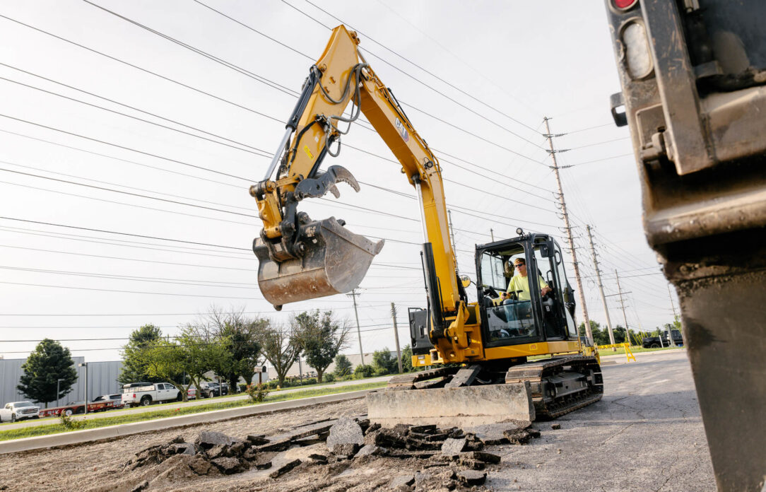 Worker uses yellow excavator on road near utility poles.