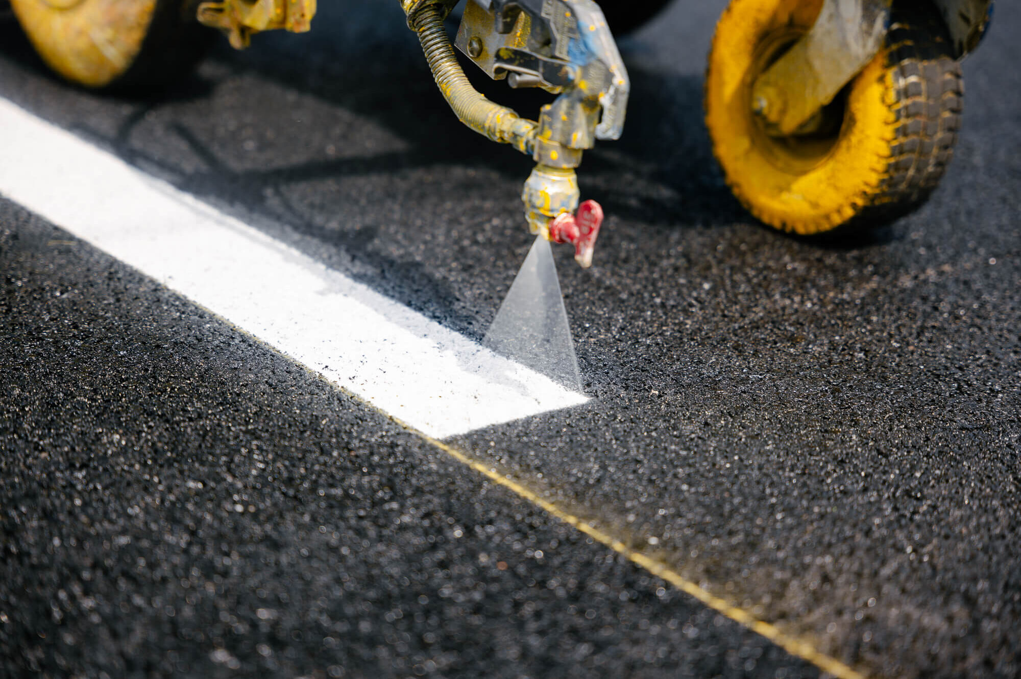 Machine paints white line on asphalt; yellow wheels behind.