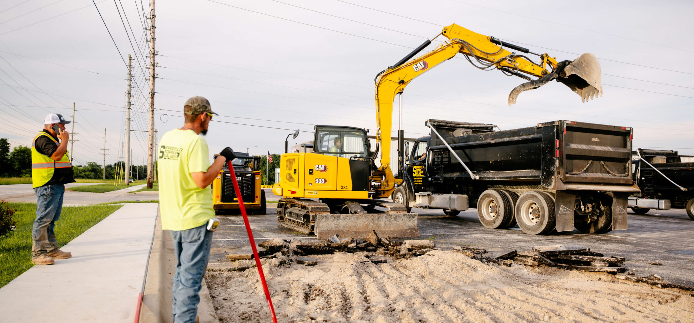 Two workers grade dirt with an excavator in the background.