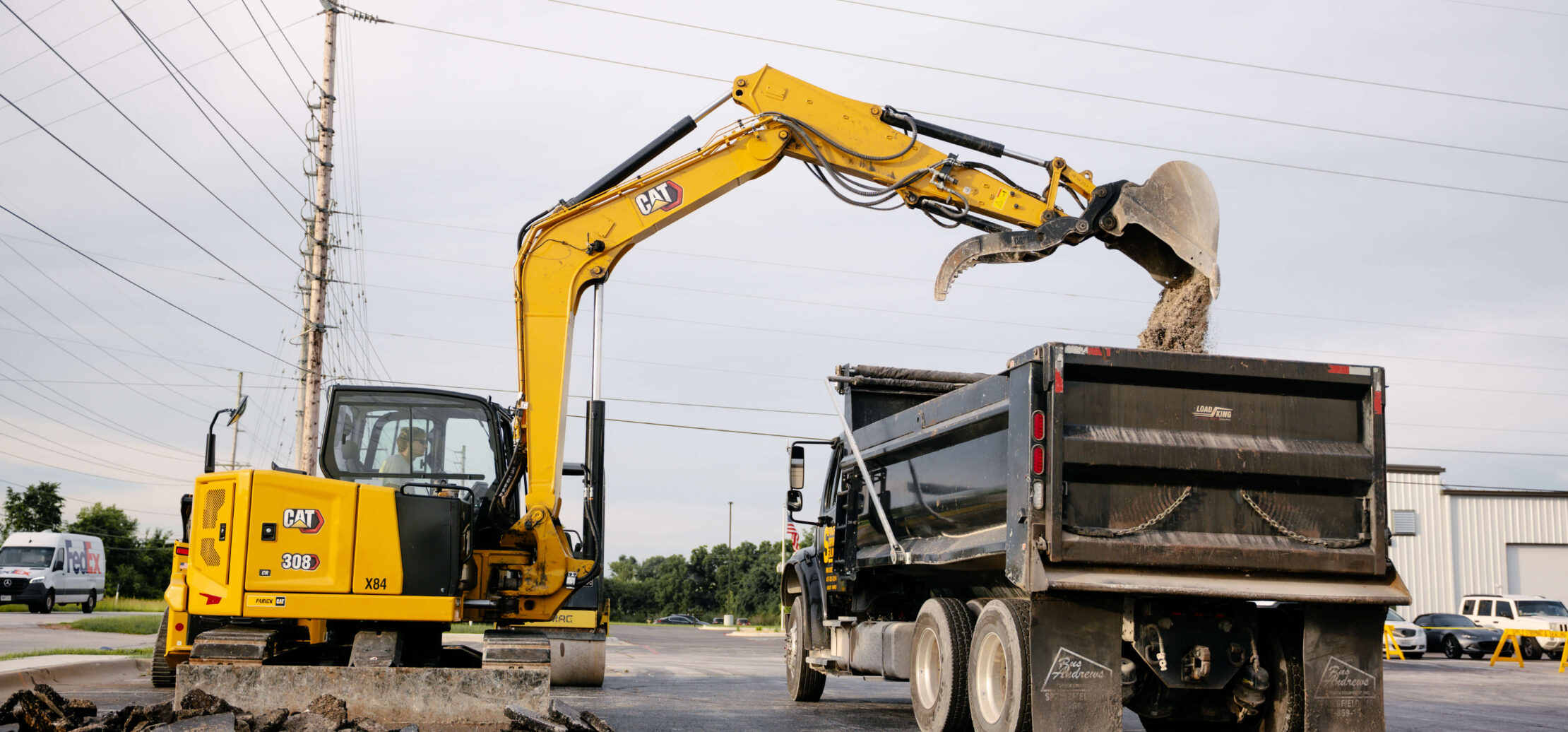 Two workers grade dirt with an excavator in the background.