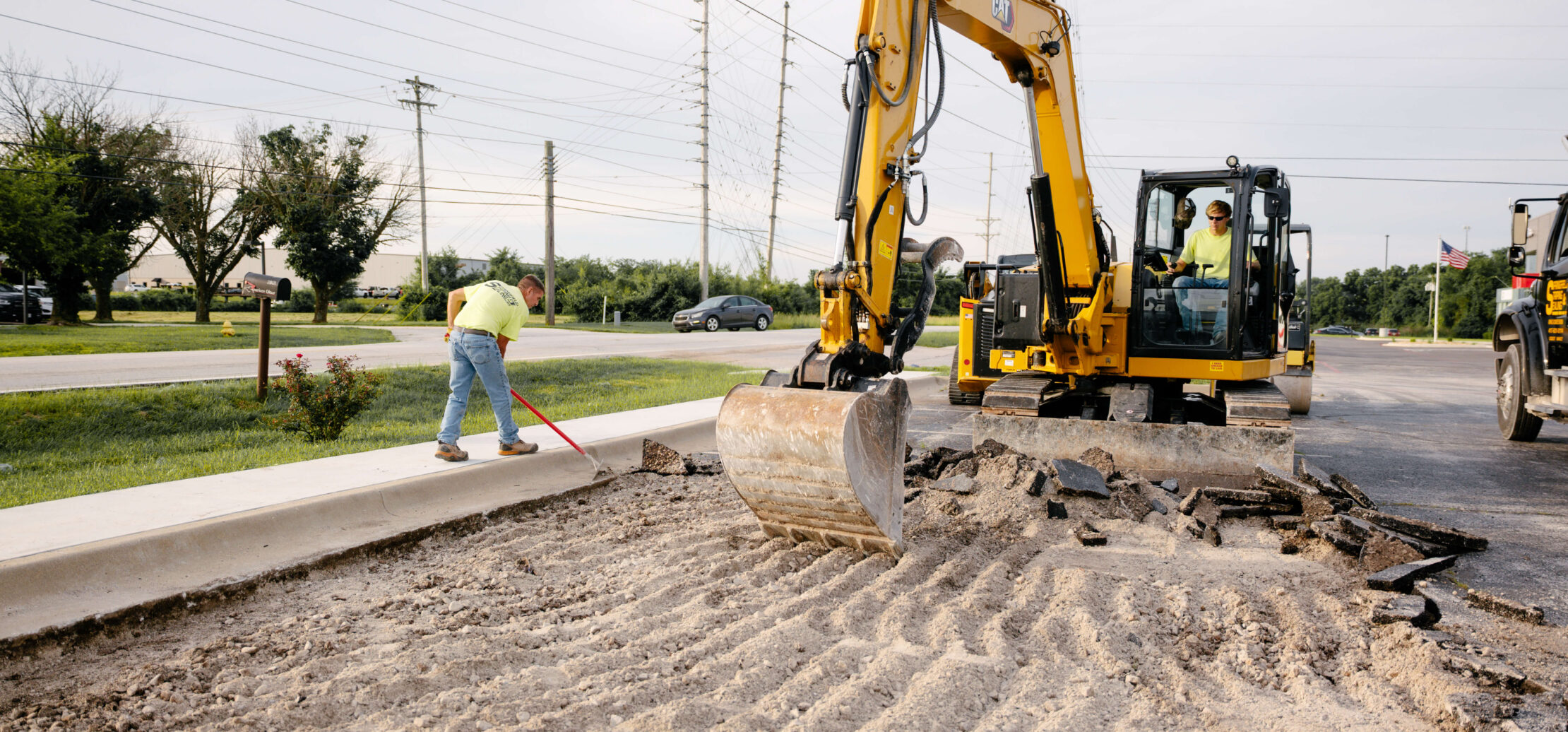 Two workers grade dirt with an excavator in the background.
