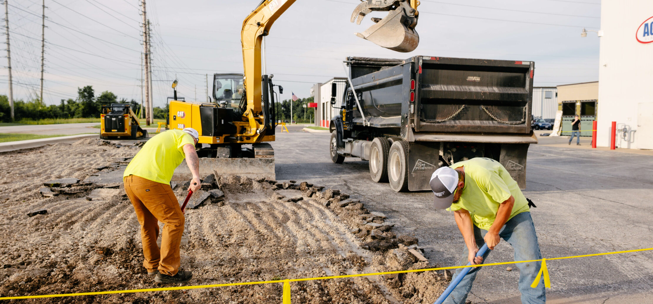 Two workers grade dirt with an excavator in the background.