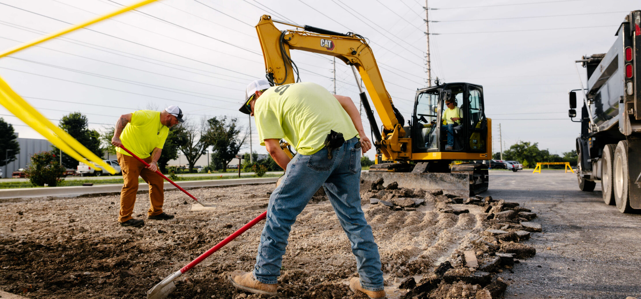 Two workers grade dirt with an excavator in the background.