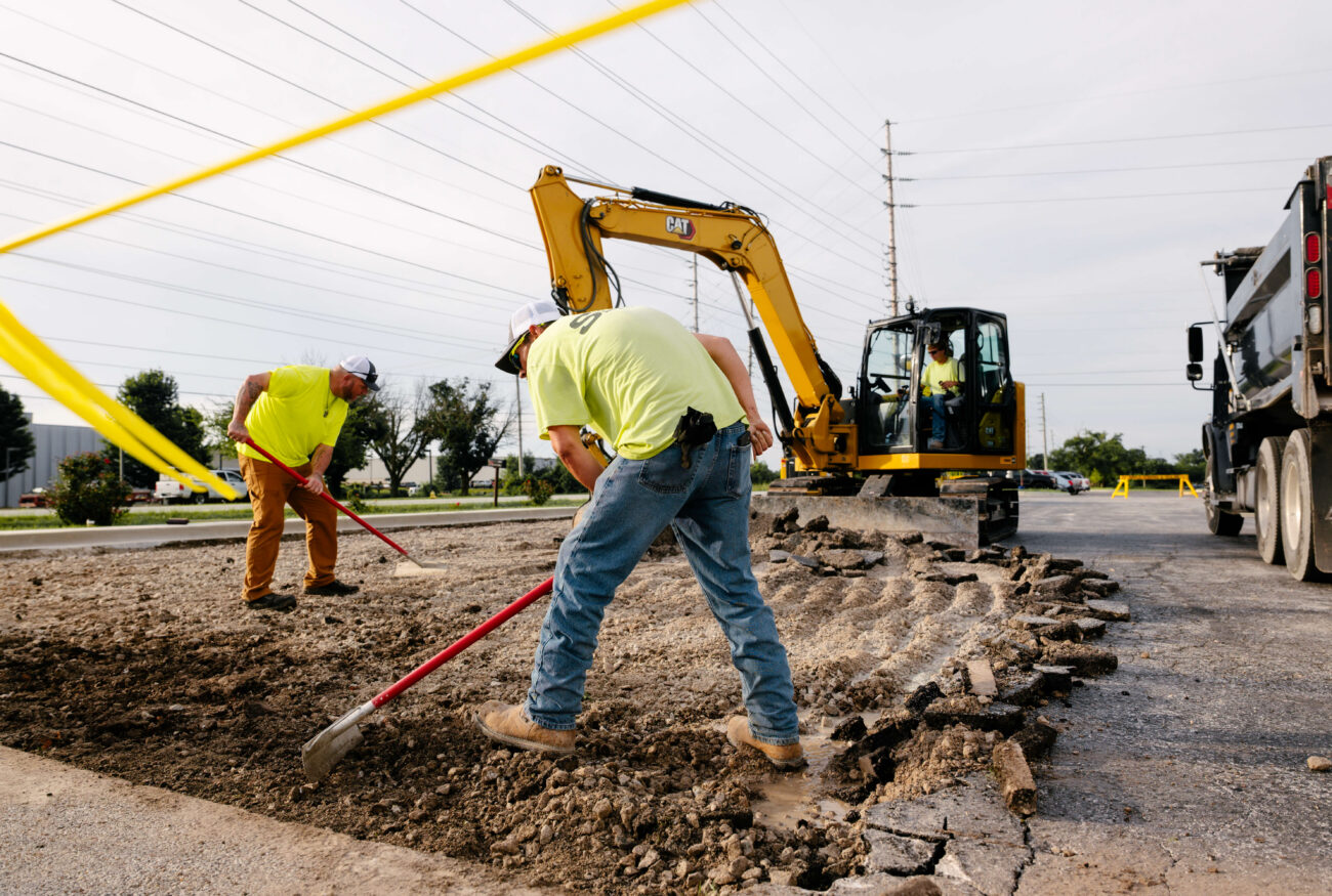 Two workers grade dirt with an excavator in the background.
