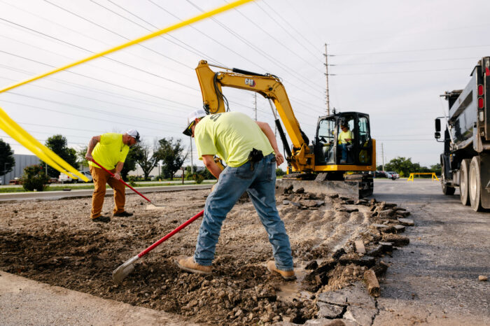 Two workers grade dirt with an excavator in the background.