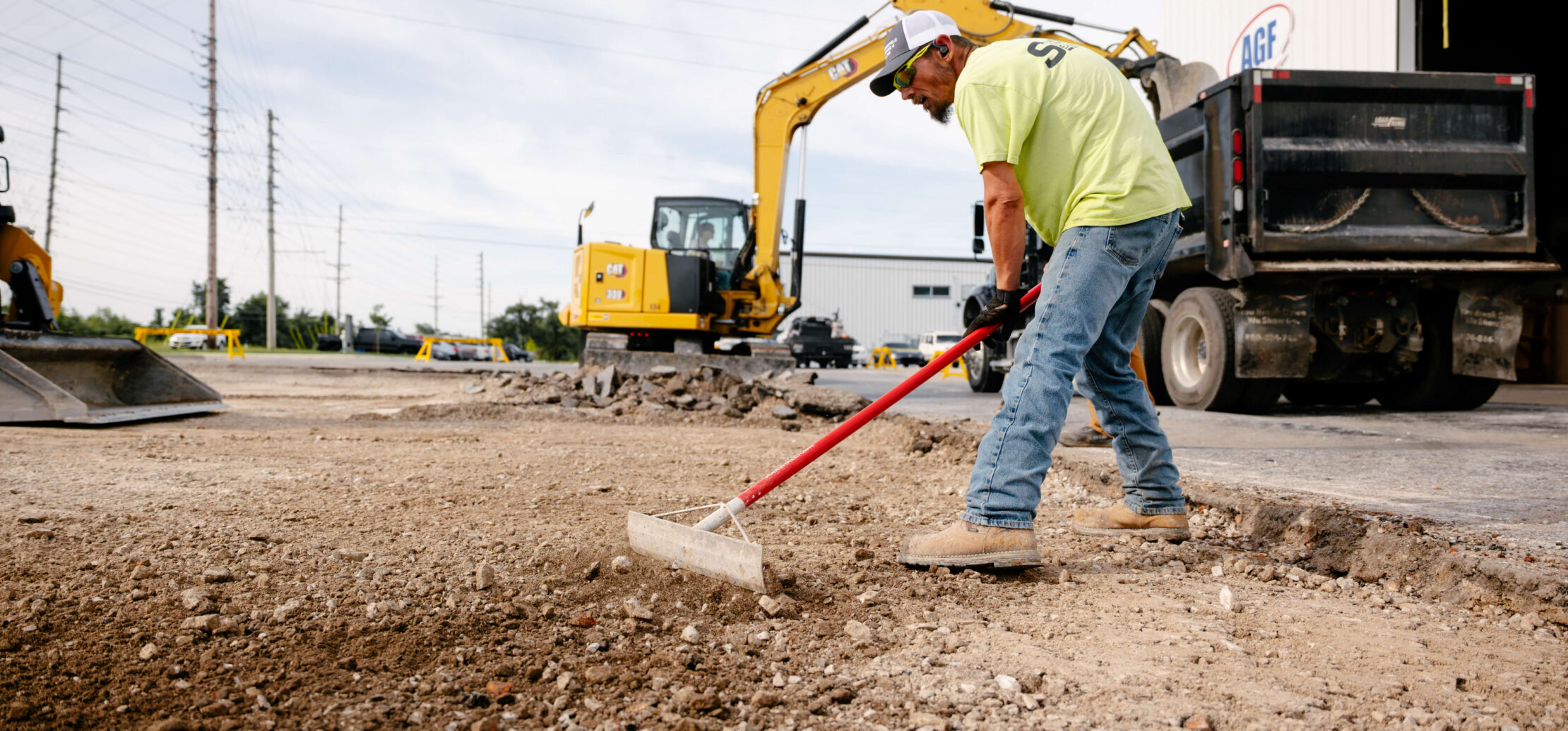 Two workers grade dirt with an excavator in the background.