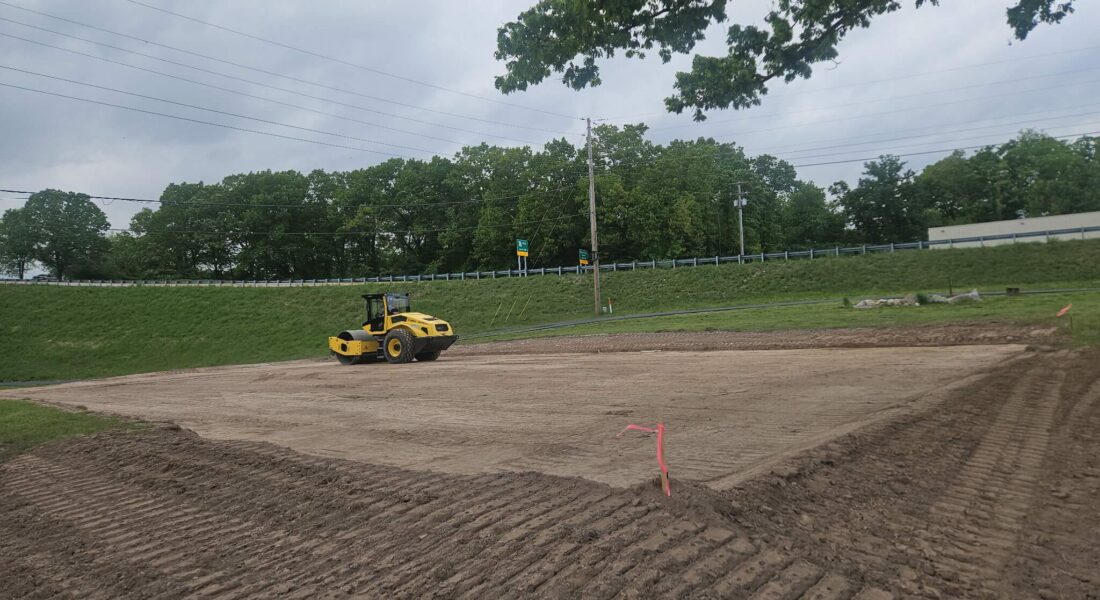 A dirt lot being flattened and leveled, gray clouds and trees in the background.