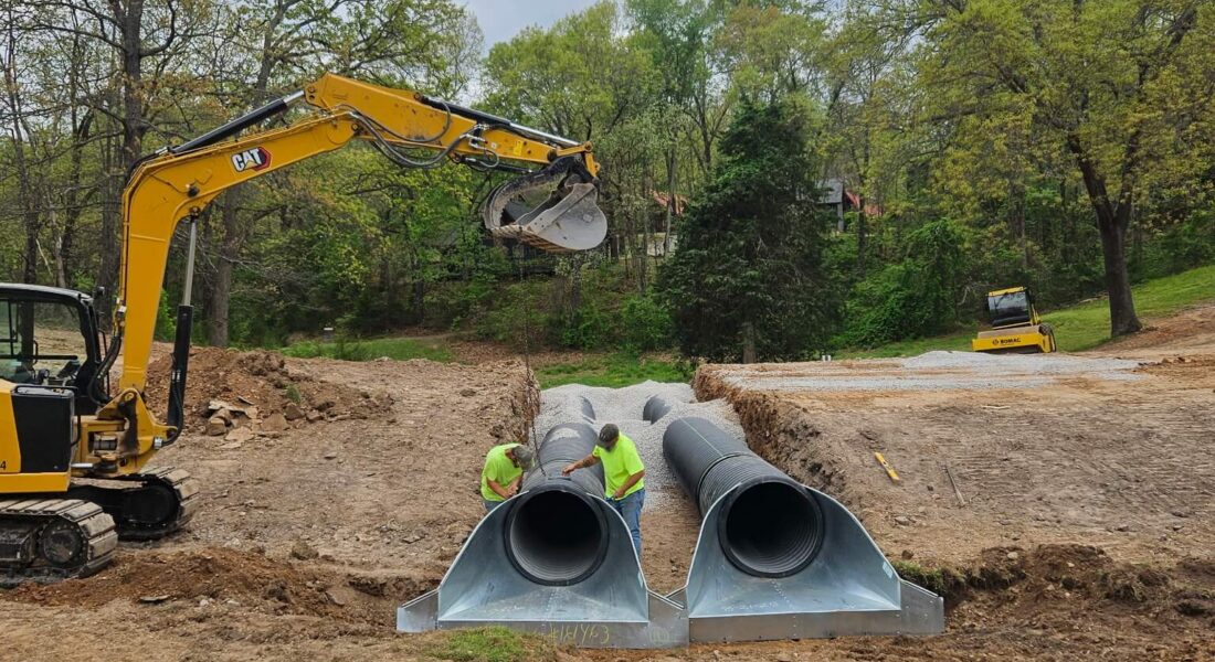 Two workers in neon shirts work on an excavation project with a yellow excavator next to them.